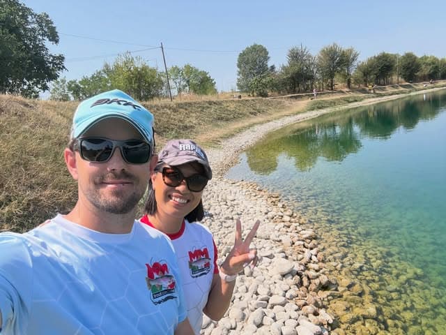 Selfie di un uomo e una donna in maglia MM, con cappellino e occhiali da sole, sulla riva sassosa di un lago