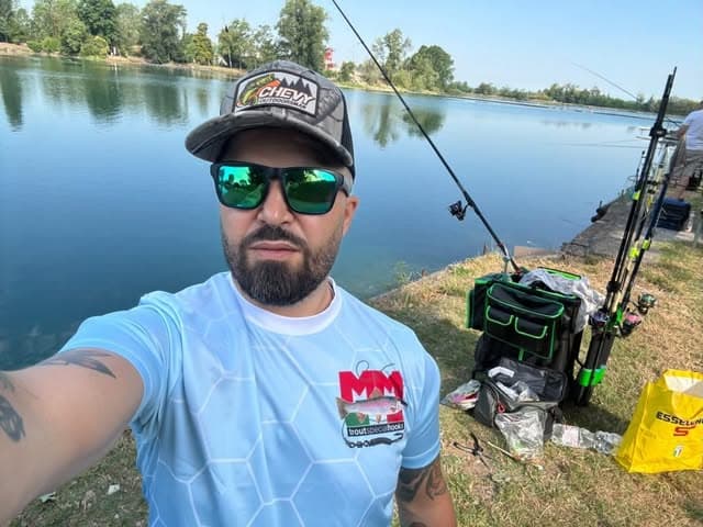 Selfie di un uomo in maglia MM davanti al lago, con le canne montate su supporto e l'attrezzatura da gara accanto